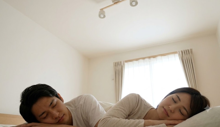 駆除が完了し、綺麗になった天井の下、明るい日差しの中で安心して眠る日本人夫婦。平穏な朝の様子。